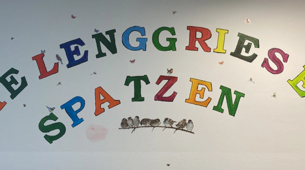 Bunter Schriftzug "Die Lenggrieser Spatzen" auf weißer Wand. 