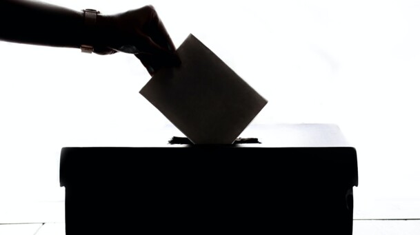 Schwarz-weiß Foto, auf dem eine Person einen geschlossenen Brief in eine Wahlurne wirft. 
