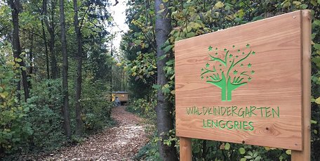 Ein braunes Holzschild mit einer hellgrünen Inschrift "Waldkindergarten Lenggries". Im Hintergrund Wald.